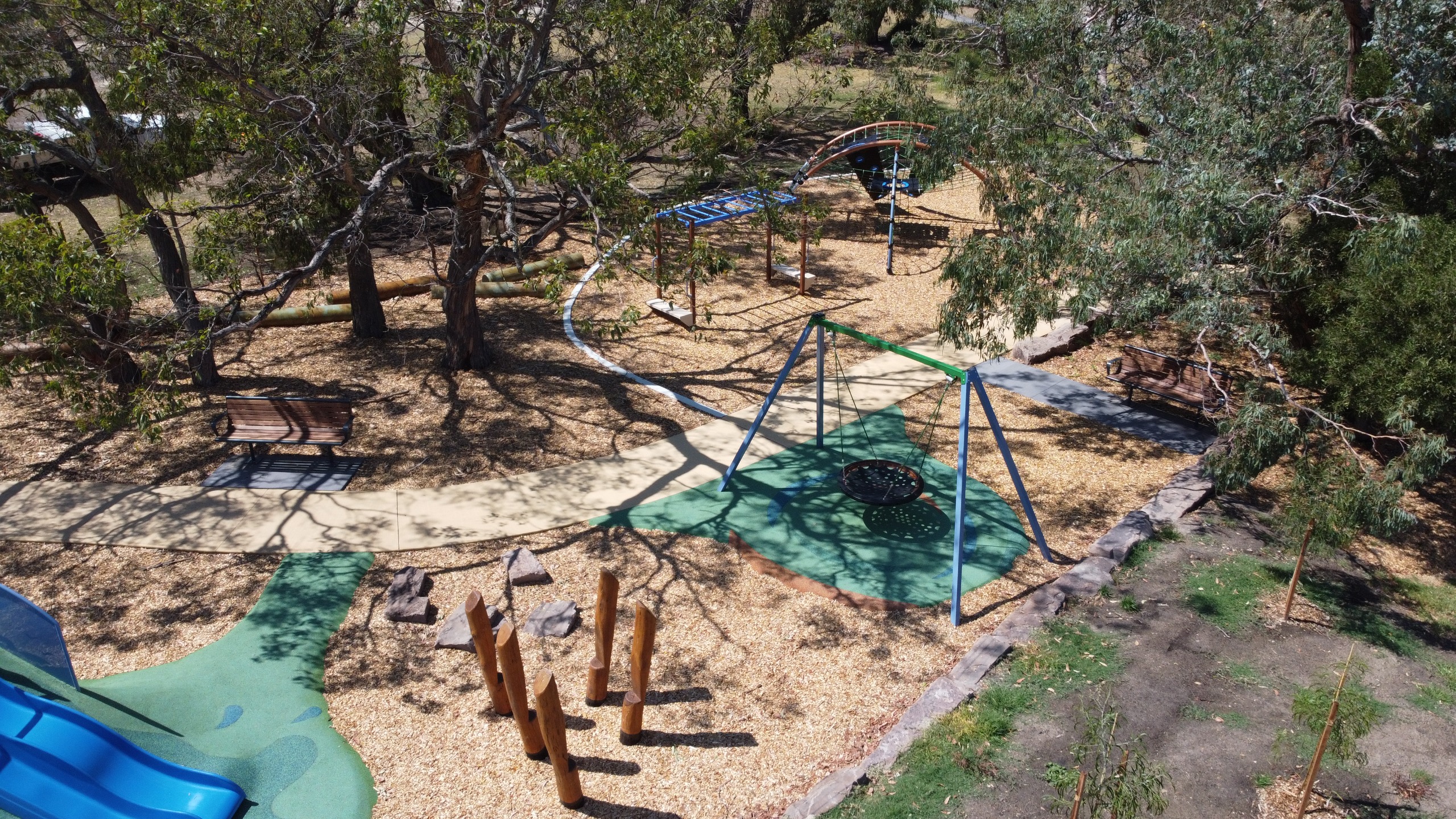 Aerial view of a playground featuring swings, climbing structures, and benches surrounded by trees and mulch.