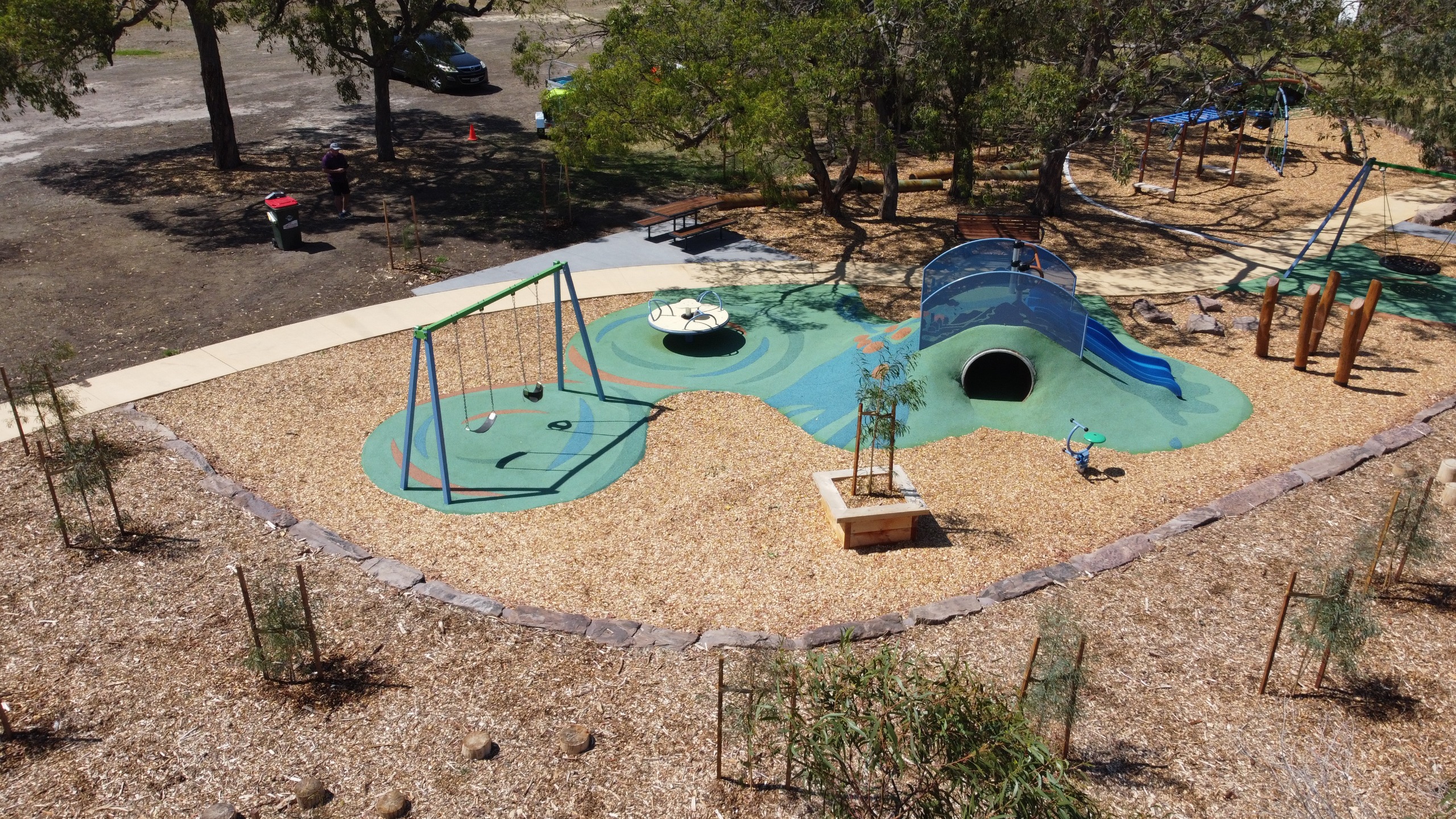A colorful playground featuring swings, a slide, and a play tunnel, surrounded by wood chips and trees.