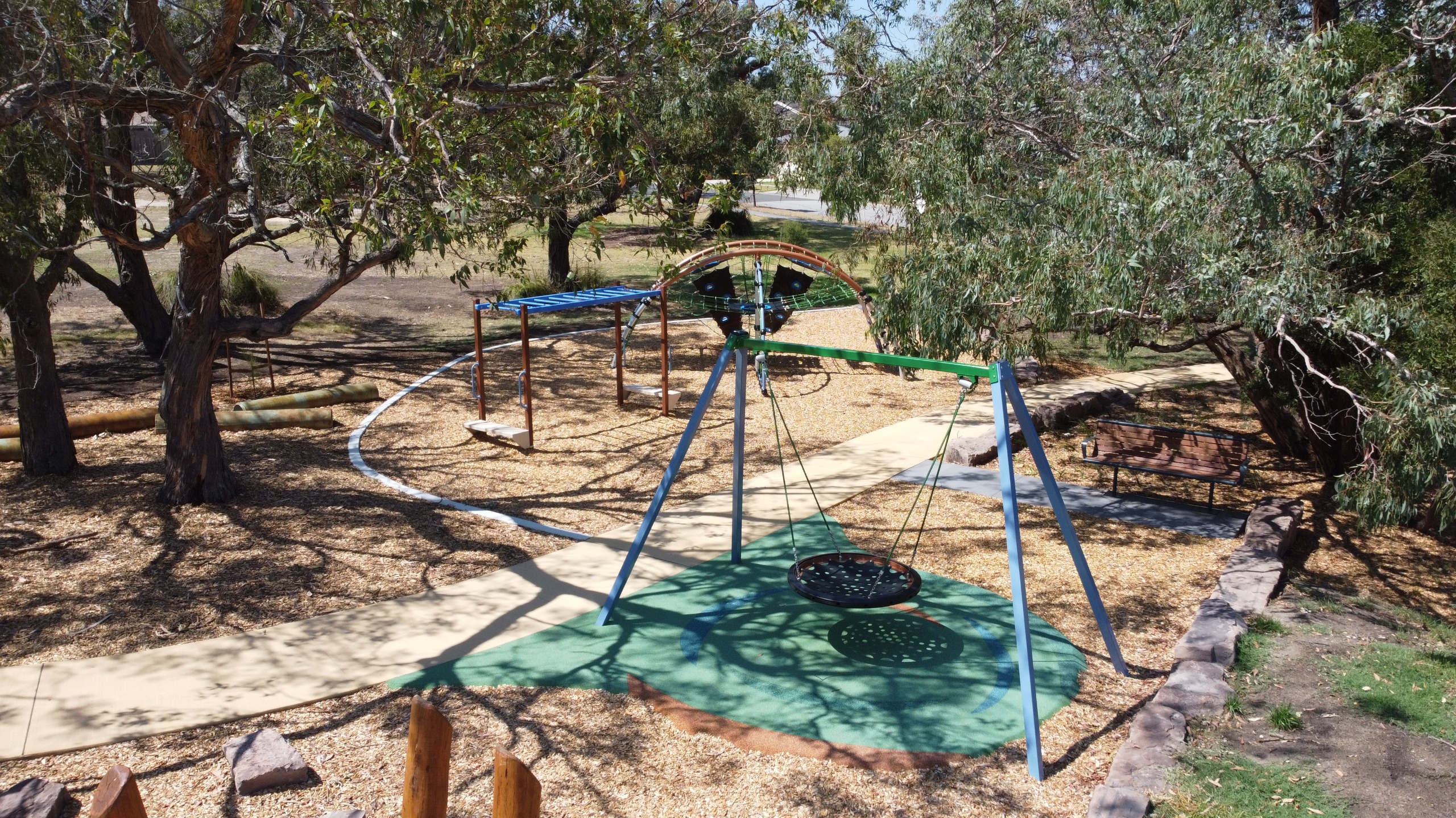 A playground scene featuring a swing set, climbing structure, and a bench, surrounded by trees and gravel pathways.