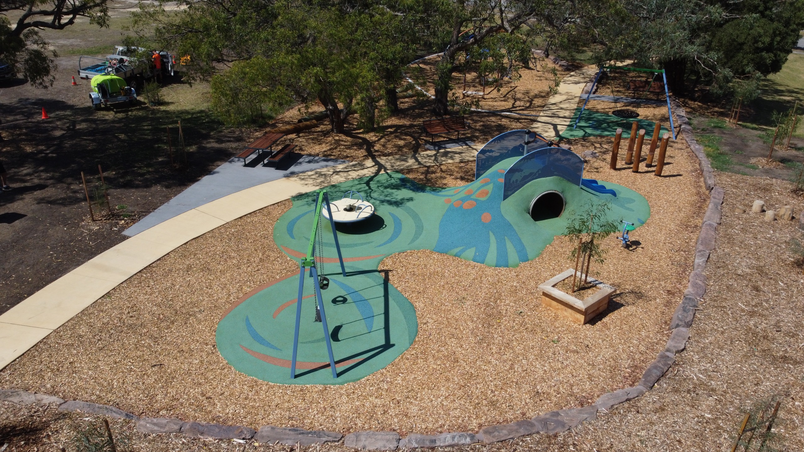 A colorful playground featuring swings, a slide, and a play structure, surrounded by mulch and trees.